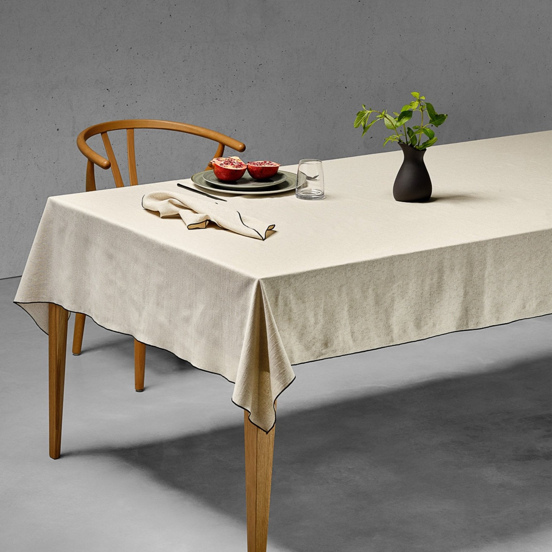 A table setting with a natural linen and cotton tablecloth (BCI cotton and Euroepean flax linen). The professionally photographed background is a concrete wall and polished concrete floor. Styled table has a handmade ceramic jug with fresh greenery, and ceramic plates. Modern mid century chair adds to the artistic style. 